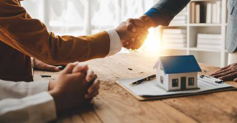 De koopovereenkomst is akkoord. 2 mensen schudden elkaar de hand met op de voorgrond een modelwoning.